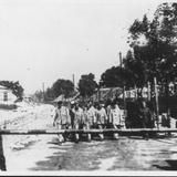 Zdjęcie archiwalne, czarno-białe. Słoneczny dzień. Grupa więźniów dochodząca do szlabanu, gdzie po bokach stoją dwaj niemieccy żołnierze. Po lewej budka i za nią budynek i wysoki komin. Po prawej drzewa zasłaniające budynki.