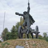 Pomnik mężczyzny stojącego na armacie z kosą. Znajduje się na niewielkim wzniesieniu porośniętym trawą.