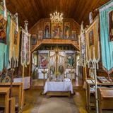 Wnętrze drewnianej cerkwi. Barwny ikonostas, ławki, chorągwie, polichromowane ściany.