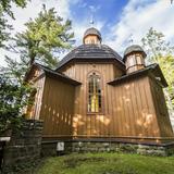 Niewielki drewniany kościółek na podmurówce z kamieni przypominający swoją architekturą małą cerkiew, widziany od dołu. Po lewej murek z kamieni i za nim wejście do kościoła z z niedużym przedsionkiem. Po prawej podobna część kościoła do przedsionka, z okrągłą wieżyczką podobną do grzyba, po środku wyższa część z wysokim, wąskim oknem i okrągłym dachem z wieżyczką. Na około drzewa i niebo częściowo zachmurzone.