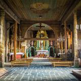 Wnętrze drewnianego kościoła. W centralnym punkcie bogato zdobiony ołtarz główny w kolorach ciemnej zieleni i złota. Na suficie widać malowidła, w nawie stoją drewniane ławki.