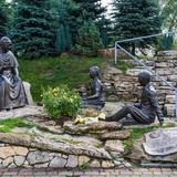 Na skałach po lewej rzeźba siedzącego św. Jana Pawła II i po środku słuchających go, siedzących dziewczyny i chłopaka. Pośrodku nich kwiaty a po prawej rzeźba leżącej kartki z napisami. Za nimi po lewej trawa i drzewa a po prawej wysoki mur z kamieni i metalowa poręcz. Za murem drzewa i mały fragment nieba.