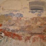 Fragment ściany synagogi z dwoma małymi oknami pod sufitem, jedno z nich zamurowane. Ściana zniszczona, przebijają na niej malowidła.