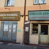 Ulica Szeroka w Krakowie. Przy ulicy znajdują się sklepy z drewnianymi okiennicami. Jeden z nich jest zamknięty. Na okiennicach wiszą plakaty a nad nimi wisi reklama 