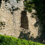 Wśród drzew liściastych widać ruiny murowanego z kamienia zamku w Lanckoronie. Fragment kamiennej ściany z łukowatym oknem z kratą.