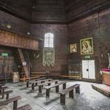 Wnętrze drewnianej kaplicy, widoczne drzwi wejściowe, ławki, obrazy..