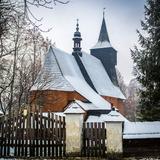 Drewniany kościół z wieżą pokryty śniegiem. Na pierwszym planie drewniana brama na murowanych słupach z żółtą tabliczką oznaczającą zabytek.