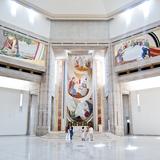 Monumentalne, jasne wnętrze sanktuarium świętego Jana Pawła, na wprost ołtarz główny, przy nim stoi kilka osób. Na ścianach kolorowe malowidła. Posadzka i sufit jasne.
