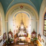 Widok z góry na wnętrze kaplicy z ostrołukowymi oknami po bokach z witrażami. Na środku w węższym od nawy prezbiterium widać ołtarz główny. Z boku ołtarze boczne. Po prawej stronie na ścianie wisi kopia cudownego obrazu Matki Bożej Sidzińskiej.