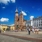 Rynek Główny w Krakowie w słoneczny dzień, w powietrzu unoszą się bańki mydlane, pośrodku w oddali widać Bazylikę Mariacką, gotycką budowlę z systemem filarowo-szkarpowym z dwiema nierównymi wieżami.