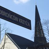 Niebieska tabliczka z nazwą ulicy: Obrońców Krzyża, w tle dzwonnica kościoła