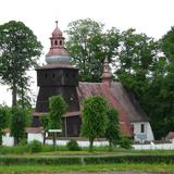 Kościół drewniano-murowany z wieżą widziany z boku, z oddali wśród drzew. Otoczony murowanym ogrodzeniem z daszkiem.