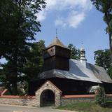 Kościół drewniany z wieżą widziany zza kaniennego ogrodzenia z bramą.