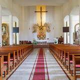 Jasne wnętrze kościoła, filary, drewniane ławki, na wprost ołtarz z Chrystusem na krzyżu i rzeźbami dwóch aniołów po bokach.