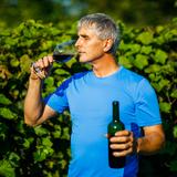 Na fotografii ujrzeć można mężczyznę ubranego podkoszulek koloru niebieskiego. W lewej ręce trzyma on butelkę z czerwonym winem, a w prawej widoczny napełniony już owym winem kieliszek. Mężczyzna degustuje wino poprzez zmysł węchu. W tle rozmyte krzewy winorośli.