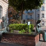 Dwie rzeźby przedstawiające postaci literatów siedzące na ławce okalającej klomb z drzewem.
