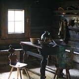 Drewniana izba z oknem i z glinianą podłogą na której stoi drewniany stół kuchenny z krzesłami. Na nim dwie misy, dzban i koszyk. Za nim półka z naczyniami i wiszącym kubrakiem.