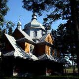 Drewniana cerkiew wśród drzew. Oświetlone dachy i kopuły, reszta budynku w cieniu.