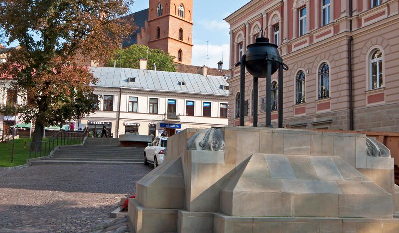 Pomnik z kamienia i płyt, zwieńczony żelaznym zniczem stoi na placu. W tle kamienice i wieża kościoła.