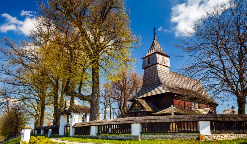 Drewniany kościół z wieżą wyrastającą z dachu, otoczony ogrodzeniem z kamienia i drewna oraz drzewami.