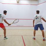 Zdjęcie przedstawia kort do gry q squasha, na którym stoi dwóch mężczyzn tyłem i grają w squasha.