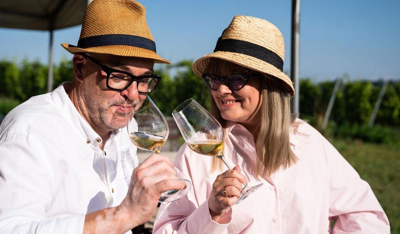 Kobieta i mężczyzna w jasnych koszulach i słomkowych kapeluszach rozkoszują się winem, w tle winnica