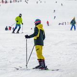 Stok narciarski ze zjeżdżającymi narciarzami, na pierwszym planie osoba w kasku z kijkami, i zapiętymi nartami ubrana w strój narciarski stoi po zjeździe