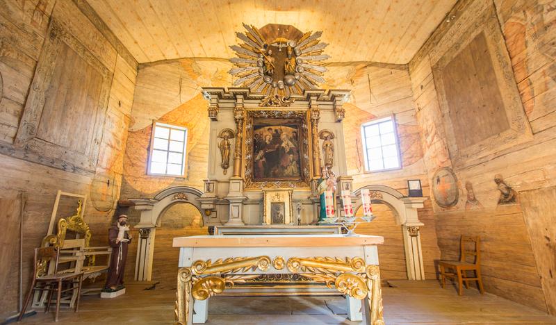 Prezbiterium w kościele. Biało-złoty ołtarz główny, drewniane ściany, pokryte częściowo malowidłami.