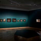 Ciemna sala wystawowa o ścianach w kolorze morskim, punktowe światło eksponuje obrazy zawieszone wzdłuż ścian. Po prawej stronie 