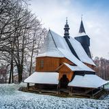 Widok zimowy. Na wprost drewniany kościół od strony zaokrąglonego prezbiterium, z podcieniami, z zadaszonym wejściem do zakrystii przybudowanej do prezbiterium. Dach stromy. Dalej wyższy dach z wieżyczką z sygnaturką na szerszej nieco nawie. Za nią widoczny górny fragment kwadratowej wysokiej wieży ze smukłą wieżyczką. Dachy zasypane śniegiem. Wokół trawnik pod cienką warstwą śniegu. i naokoło wysokie drzewa bez liści. Niebo częściowo zachmurzone.