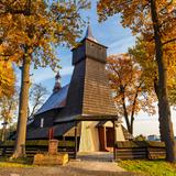 Drewniany kościół z wysoką wieżą otoczony drewnianym ogrodzeniem oraz kilkoma drzewami o żółtych i pomarańczowych liściach.