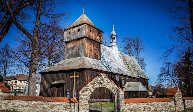 Drewniany prosty kościół z niską wieżą, otoczony kamiennym ogrodzeniem.