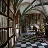 Biblioteka klasztorna, posadzka w szachownicę, po prawej fotel i inne drewniane, rzeźbione meble. Po lewej i na wprost wysokie regały pełne starych ksiąg. Freski na żebrowanym suficie.