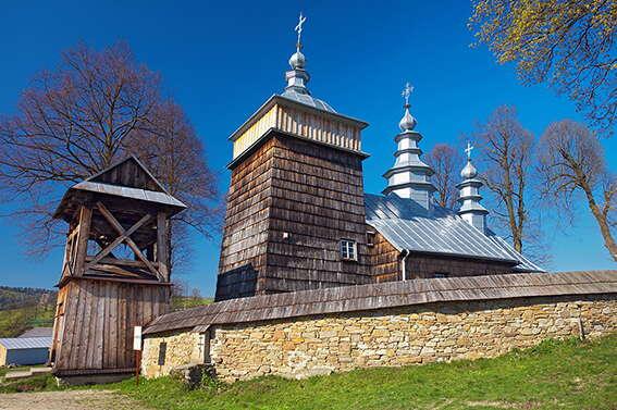 Drewniana cerkiew zza murowanego ogrodzenia z dzwonnicą.