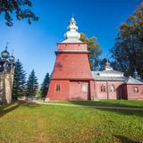 Drewniana cerkiew o ścianach w kolorze czerwonobrązowym i jasnym dachu. Przed nią kamienna dzwonnica.