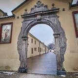 Pięknie zdobiona brama klasztorna w ciemnym obramowaniu. Po bokach na murze wiszą obrazy świętych. Przez bramę widać zabudowania klasztorne.