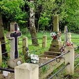Krzyż, pomnik i mogiły na cmentarzu, z tyłu ogrodzenie, dużo zieleni.