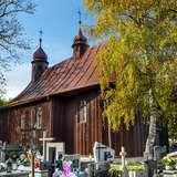Drewniany kościół z blaszanym dachem i dwoma wieżyczkami na sygnaturkę, stojący na cmentarzu. Wokół nagrobki i drzewa.