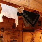 Wnętrze muzeum Willa Koliba, drewniane pomieszczenie, ściany wykonane z drewnianych desek, na ścianach wiszą eksponaty i tradycyjne góralskie przyrządy. U sufitu na belce drewnianej wisi biała koszula i kolorowa chusta.