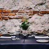 Zbliżenie na stolik w Restauracji Amarylis w Krakowie. Na nim czarny obrus, a z kolei na nim szklanki, kieliszki, serwetki, kwiat  i świeczka. Za stołem ceglano-kamienna ściana,
