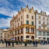 Na wprost za dużym placem ze spacerującymi ludźmi bogato zdobiona czterokondygnacyjna kamienica. Na parterze duże okna z łukami, nad oknami pierwszego piętra zdobienia i łuki. Wyżej mniejsze, prostokątne okna. U góry dach zasłania zdobiona, falista attyka. Za kamienicą ciągną się inne, niższe. Po lewej fragment ceglanego kościoła. Po prawej wysoka, druga kamienica. Za nią dalej kolejna, równie wysoka. Pod kamienicami stoją parasole. Na niebie cienkie chmury.