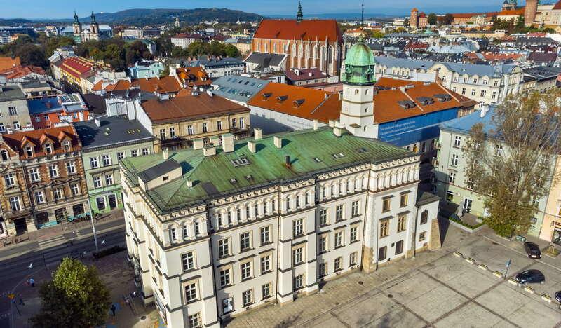 Biały budynek z zielonym dachem należący do Muzeum Etnograficznego w Krakowie na Kazimierzu. Przed nim znajduje się pusty plac, a w oddali widać kamienice, kościoły i inne budynki.