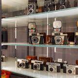 Stare aparaty fotograficzne poukładane na trzech szklanych półkach w szklanej gablocie w Muzeum Fotografii w Krakowie. Za aparatami znajdują się skórzane etui, a aparaty na drugiej półce mają też duże lampy błyskowe.