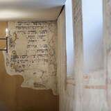 Wnętrze synagogi Kupa w Krakowie. Przybliżenie na ścianę z napisami po hebrajsku. Obok ściana również z napisami i malunkami. U sufitu wisi metalowy żyrandol z żarówkami w formie świec.