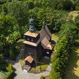 Panorama z lotu ptaka - drewniany kościół z kwadratową wieżą z przodu, w otoczeniu drzew i zieleni.