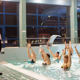 Instruktorka pokazująca ćwiczenia na zewnątrz basenu, uczestniczki ćwiczące w basenie.