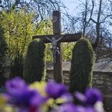 Drewniany krzyż z ukrzyżowanym Chrystusem stojący między dwoma ładnie przyciętymi, ukształtowanymi tujami. Za nim fragment ogrodzenia z kamienia. Przed nim zamglone fioletowe i żółte kwiaty.