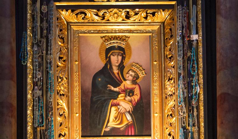 Obraz Madonny z Dzieciątkiem w złotej ramie. Wizerunek przykryty sukienkami i koronowany. Ołtarz czarny, złocony.