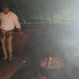 Mężczyzna w stroju góralskim stojący pod ścianą wewnątrz drewnianego szałasu. Nad ogniskiem wisi miedziane naczynie z mlekiem.